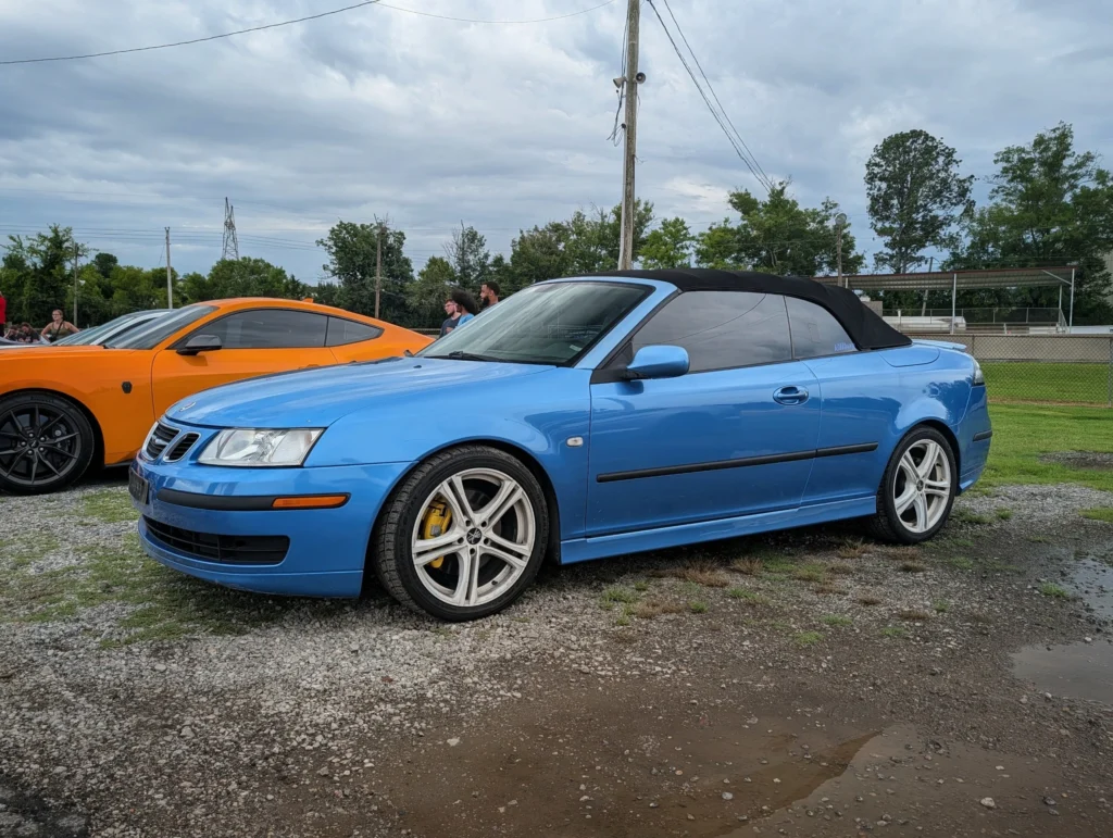 Blue Saab 9-3 Aero and orange Ford Mustang parked side by side at an outdoor car event with people and trees in the backgr