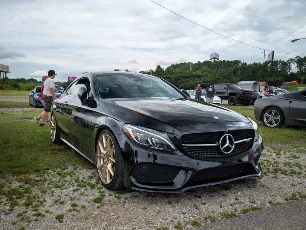 Black Mercedes-Benz AMG C-Class with gold wheels parked on gravel beside two pedestrians and various other vehicles under overcast