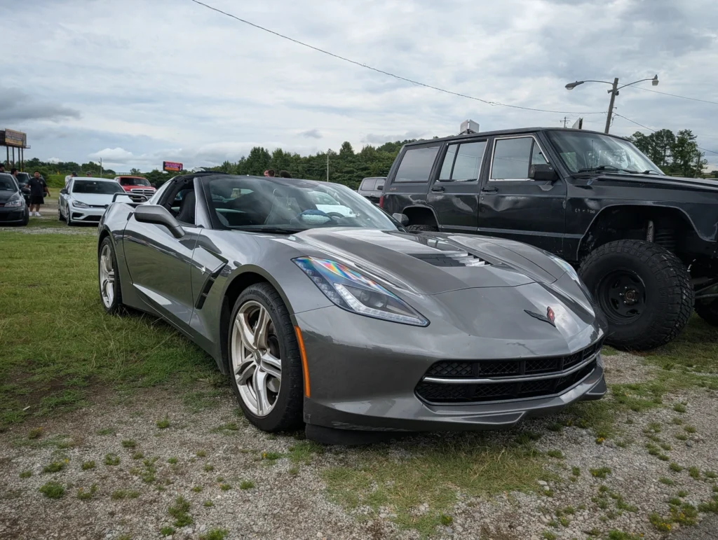 Gray Chevrolet Corvette convertible parked on grass with other vehicles and people in the background under a cloudy sky