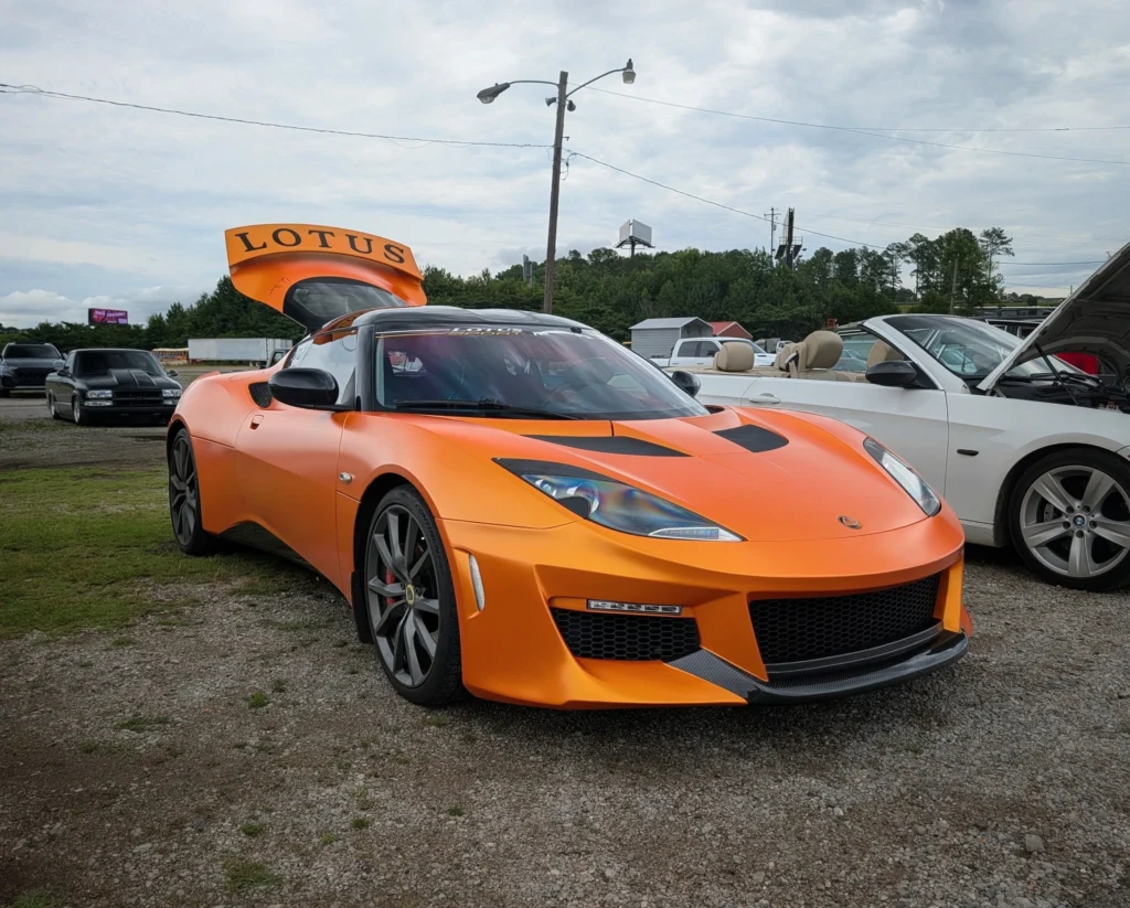 Orange Lotus Evora GT with open rear hatch parked on gravel with other vehicles and trees in the background