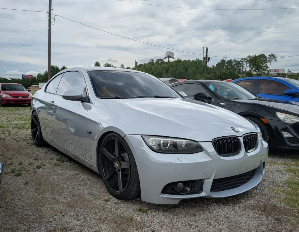 Silver BMW 3 Series parked on gravel with other cars in background under cloudy sky