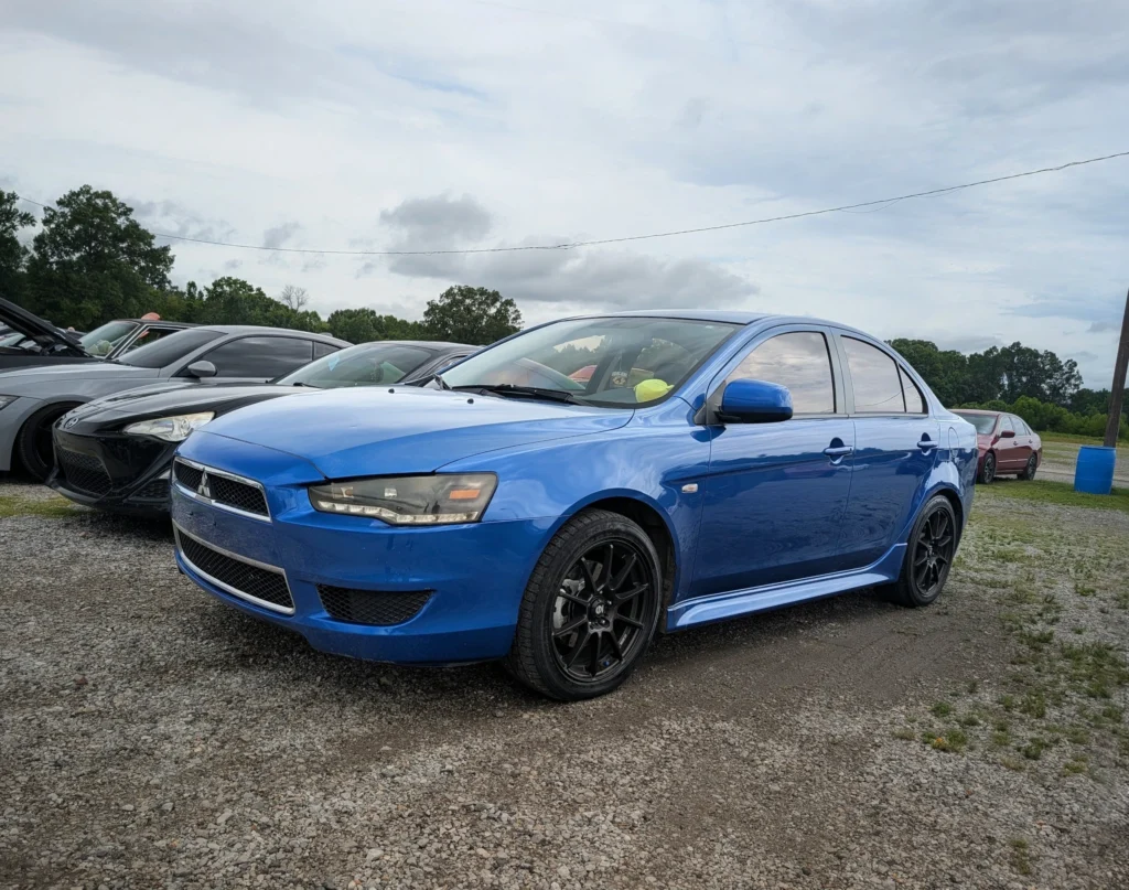 Blue Mitsubishi Lancer parked on gravel with other cars at an outdoor event under cloudy sky