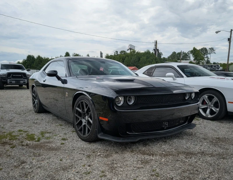 Black Dodge Challenger parked on gravel alongside a white Dodge Challenger and a white RAM truck under a cloudy sky.