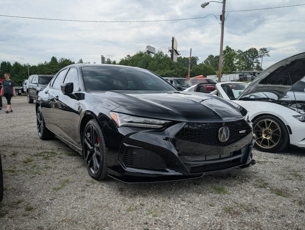 Black Acura TLX parked on gravel lot at car event with white convertible next to it and people walking nearby
