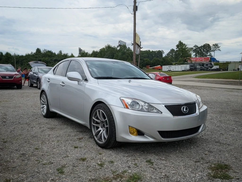 Silver Lexus IS sedan parked on gravel with other vehicles and people in a parking lot under cloudy sky.