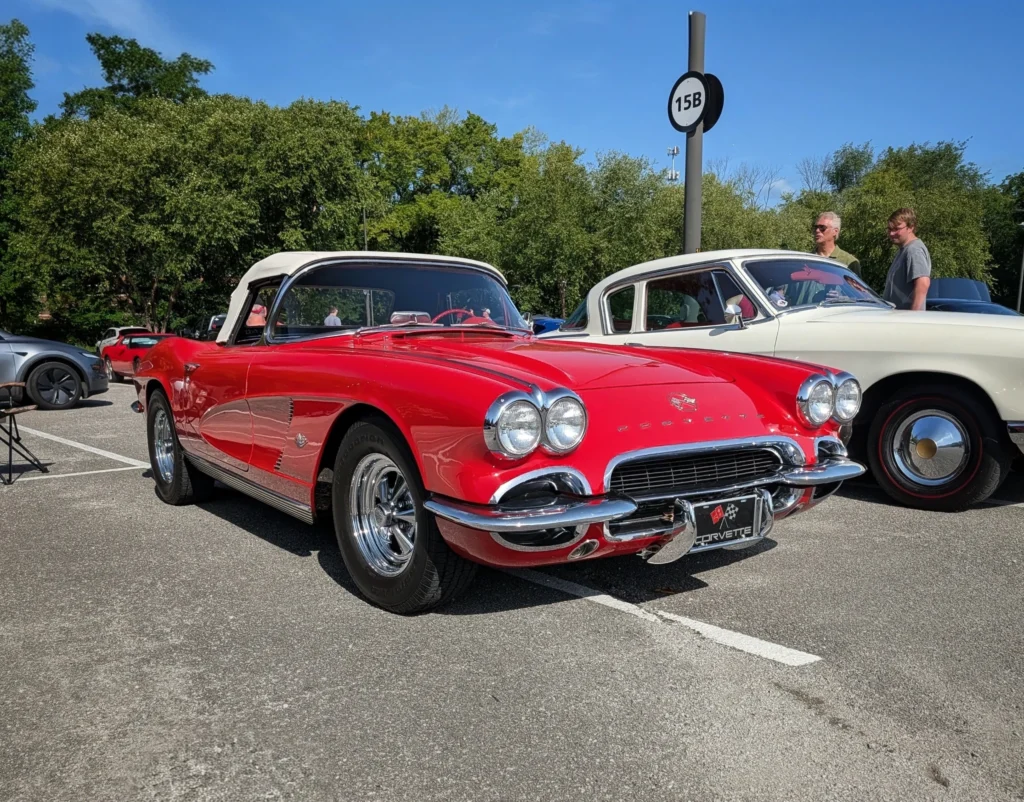 Red Chevrolet Corvette convertible with white top parked next to a white coupe in a sunny outdoor lot with tall trees backgro