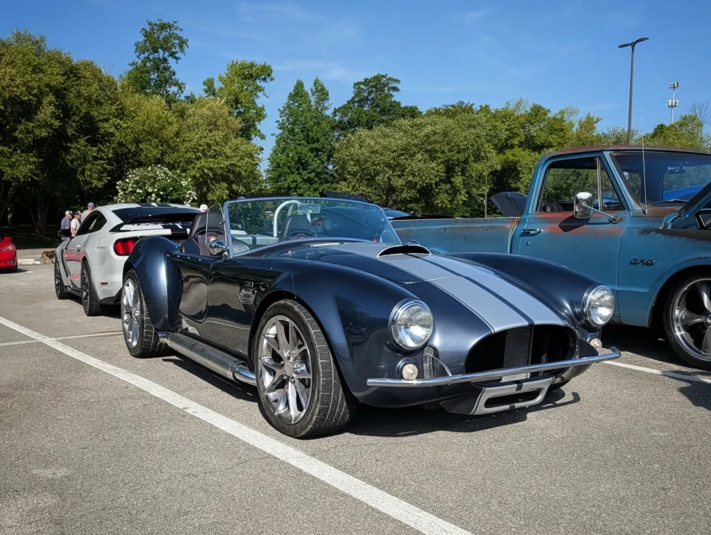 Black AC Cobra with silver racing stripes parked beside a white Ford Mustang and a blue GMC pickup in an outdoor lot with tre