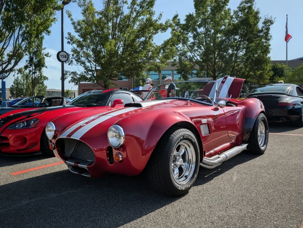 Red Shelby Cobra replica roadster with white racing stripes parked at a sunny outdoor car show