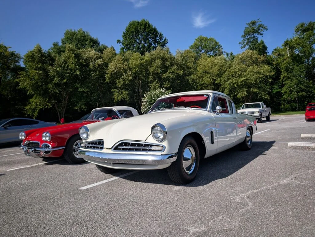 White Studebaker car parked next to a red convertible and other vehicles in a lot with green trees in the background