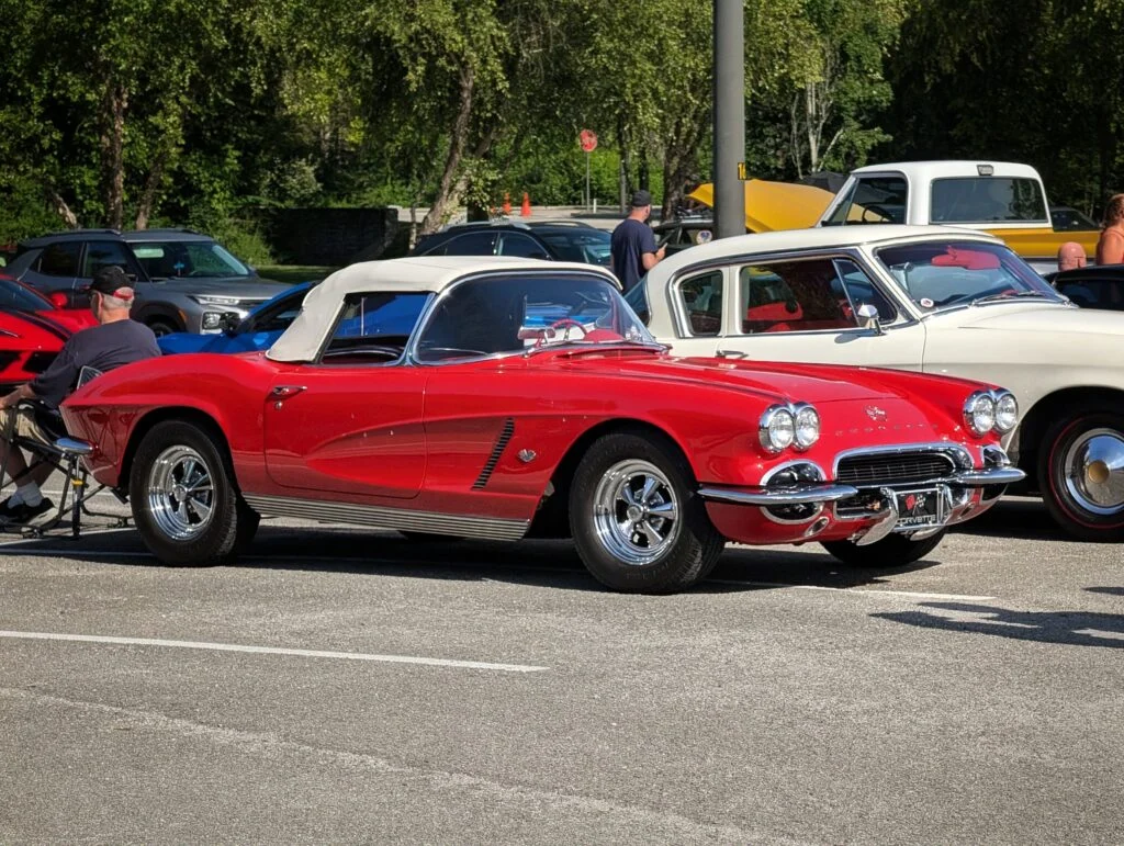 Red Chevrolet Corvette convertible with white soft top parked among other classic cars and attendees at an outdoor car event