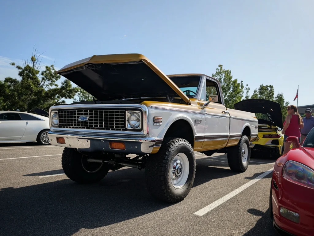 White and gold Chevrolet pickup truck with hood open at an outdoor car event with other cars and people in background.
