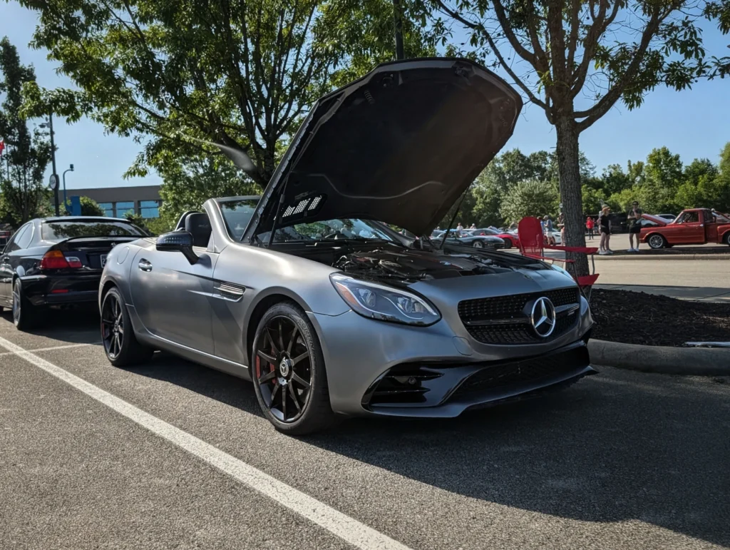 Silver Mercedes-Benz AMG SLC 43 with hood open parked at an outdoor car event with other vehicles and people in background