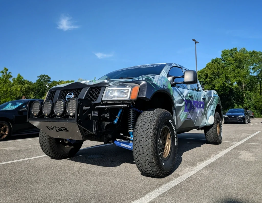 Camouflage patterned Nissan truck with off-road lights on grill in a parking lot with trees in background