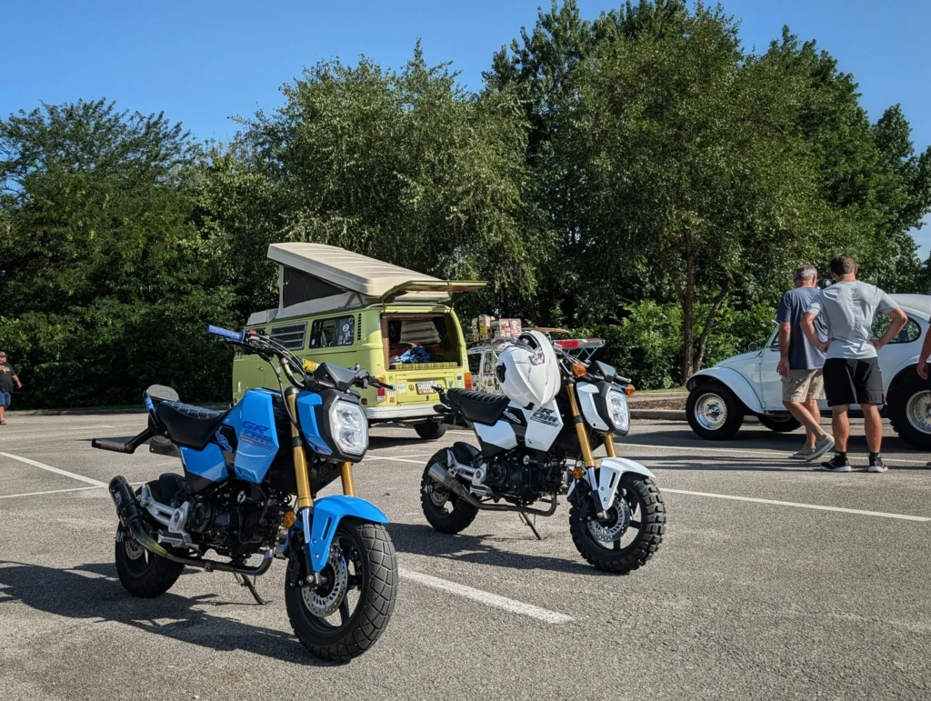 Blue and white Honda Grom motorcycles parked in front of a yellow Volkswagen camper van with its roof popped up and people ne