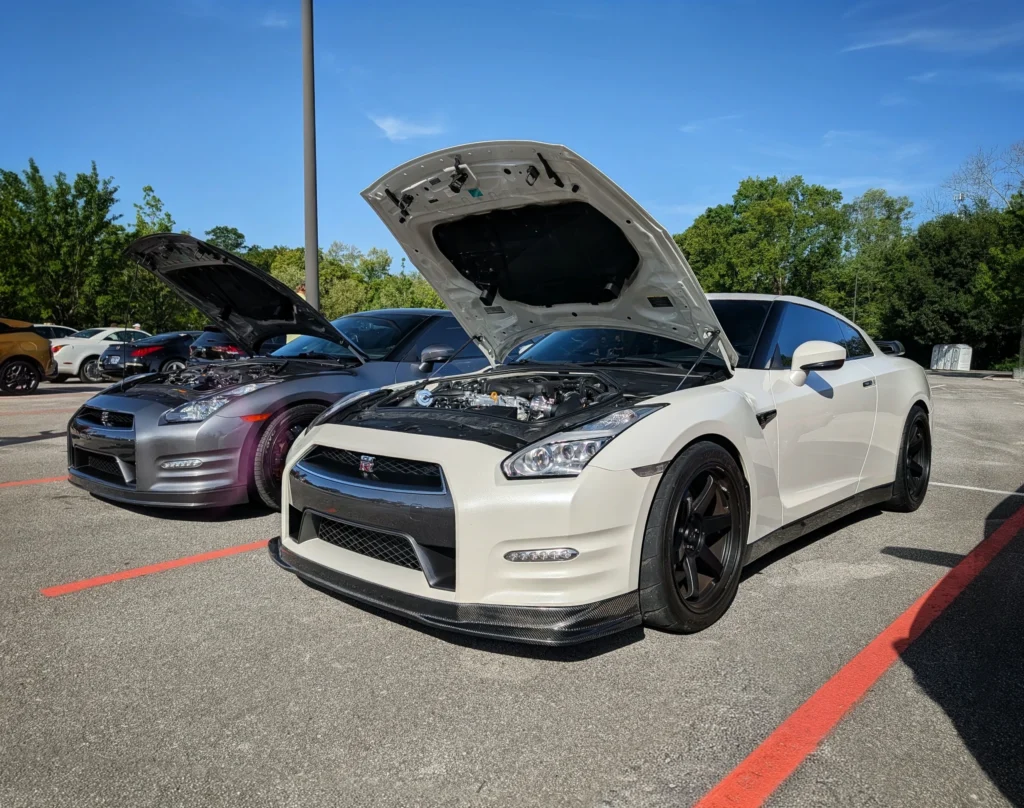 White Nissan GT-R and silver Nissan GT-R parked side by side with hoods open in a parking lot during daytime
