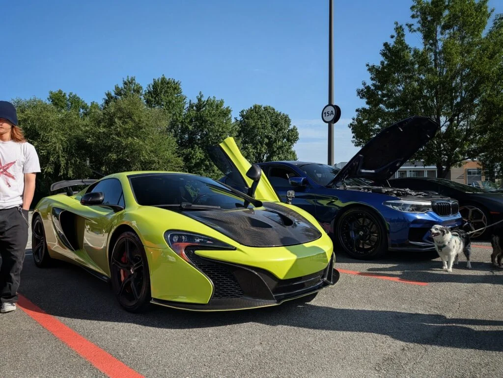 Yellow McLaren with butterfly doors open at outdoor car event with blue BMW and trees in background