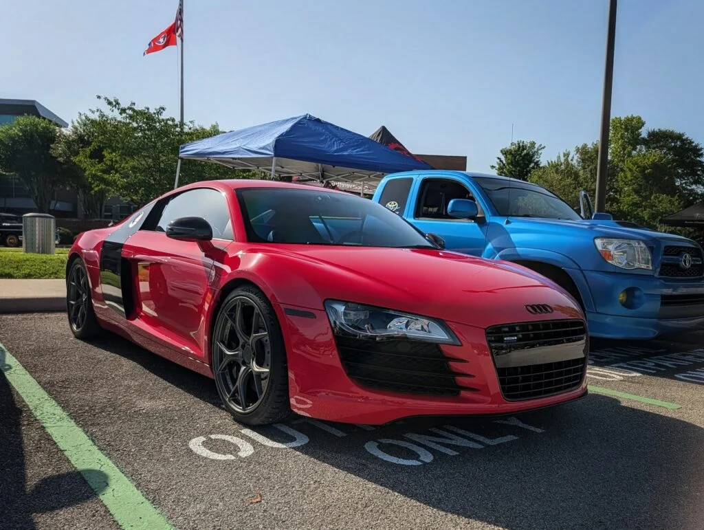 Red Audi R8 and blue Toyota Tacoma parked side by side at an outdoor car event with tents and trees in background