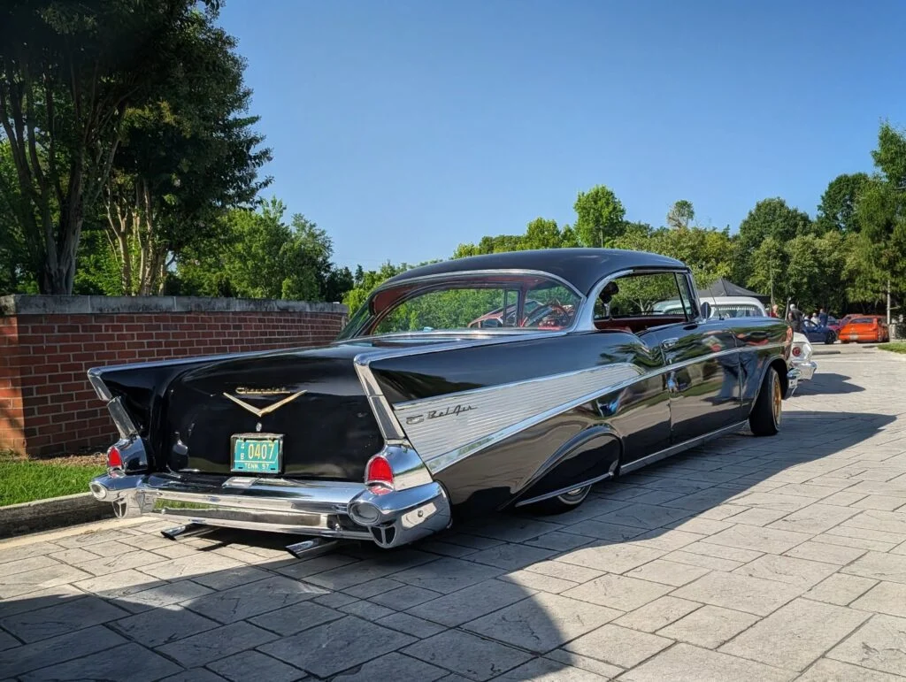 Black 1957 Chevrolet Bel Air parked outdoors near a brick wall with trees and other vehicles in the background