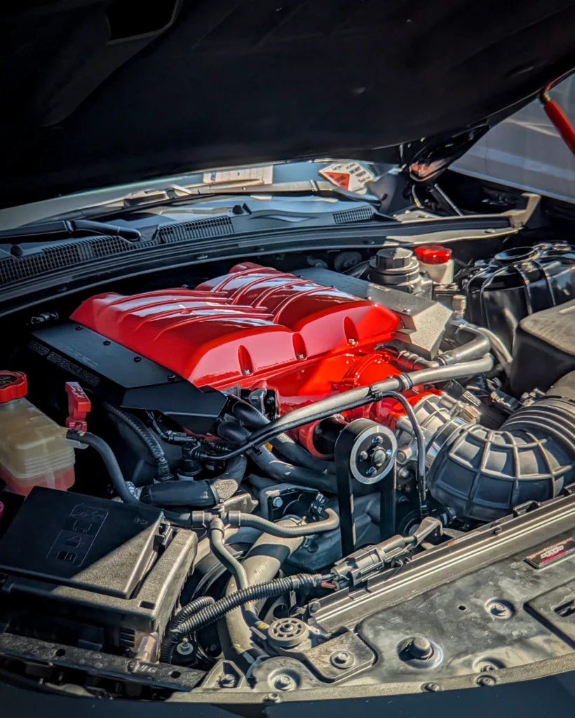 Close-up of a car engine bay with a red intake manifold and various engine components under a lifted hood.