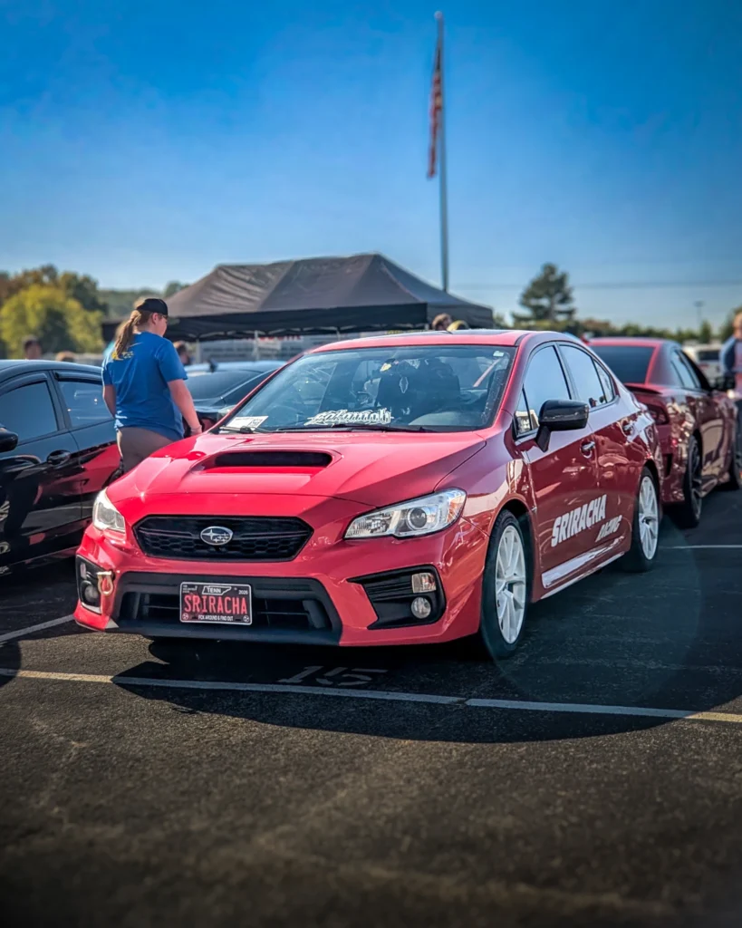 A red Subaru WRX with white wheels is parked at an outdoor car show beside other cars and attendees.