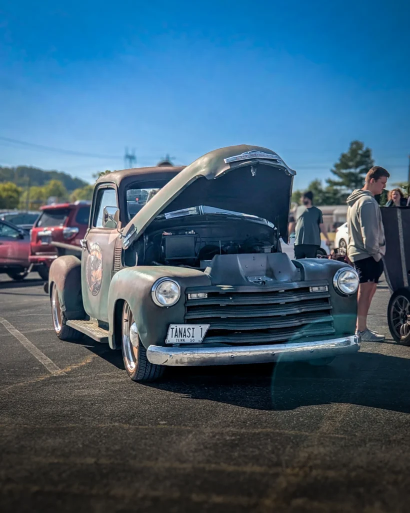 A weathered green Chevrolet 3100 pickup with its hood raised sits in a parking lot with attendees and other vehicles.