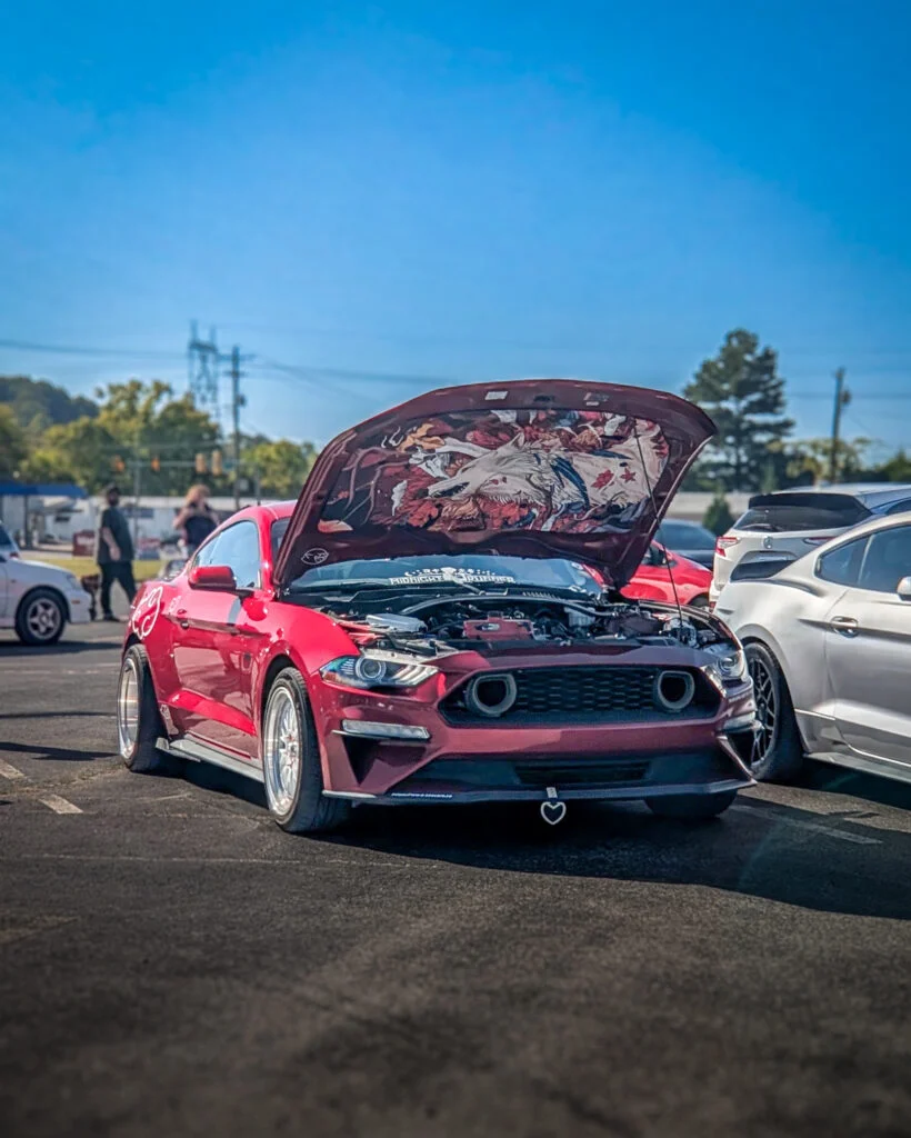 Red Ford Mustang with hood open showing custom artwork on underside, parked at an outdoor event with people and cars nearby