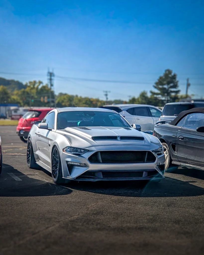 Silver Ford Mustang parked in a lot with other cars under a clear blue sky