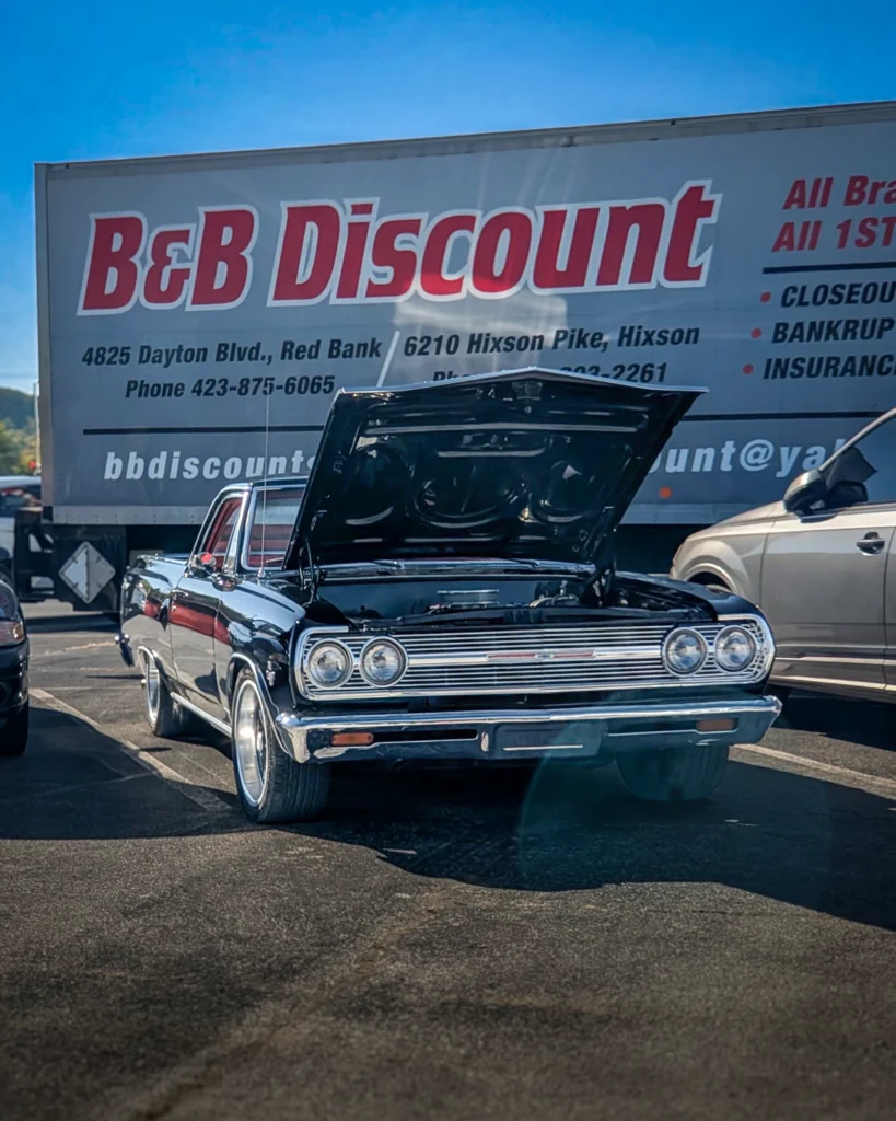 Black Chevrolet Impala with hood open at an outdoor event parking lot in front of a B&B Discount truck
