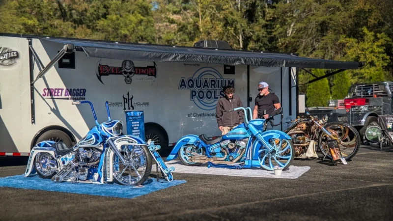 Two men standing near custom blue motorcycles parked in front of a trailer with advertising, outdoors on a sunny day