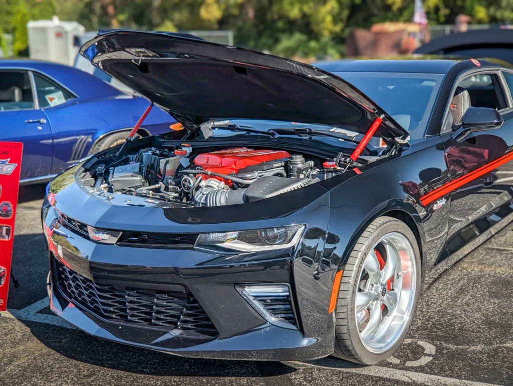 Black Chevrolet Camaro with hood open showing engine at a car meet with other vehicles in the background