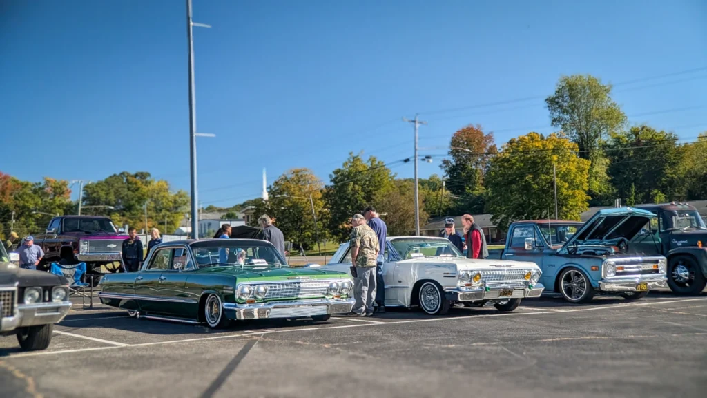 Several vintage Chevrolet vehicles in a parking lot with people standing around on a clear day with blue sky and trees in