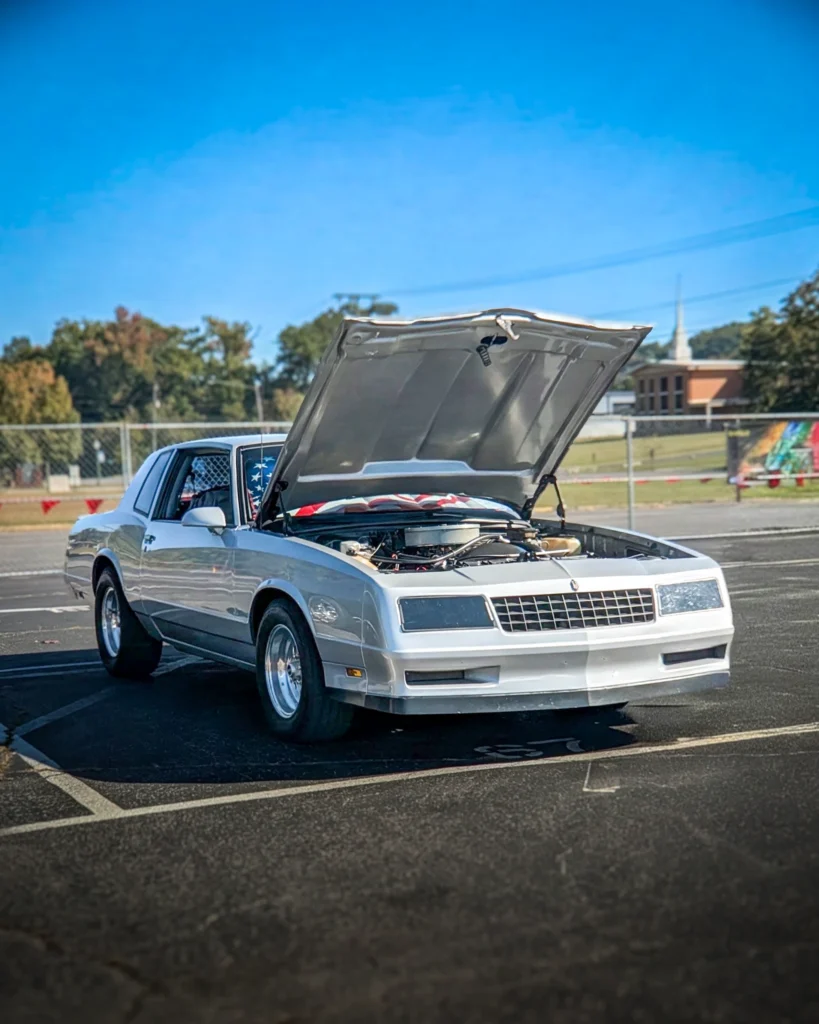 White Chevrolet Monte Carlo with hood open in parking lot at a car event under clear blue sky