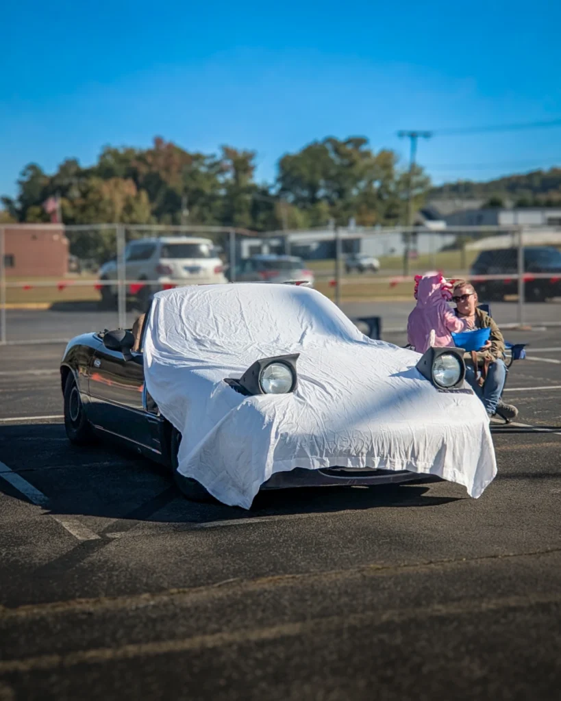 Mazda Miata with a white cover over the front, pop-up headlights visible, parked outdoors with two people sitting