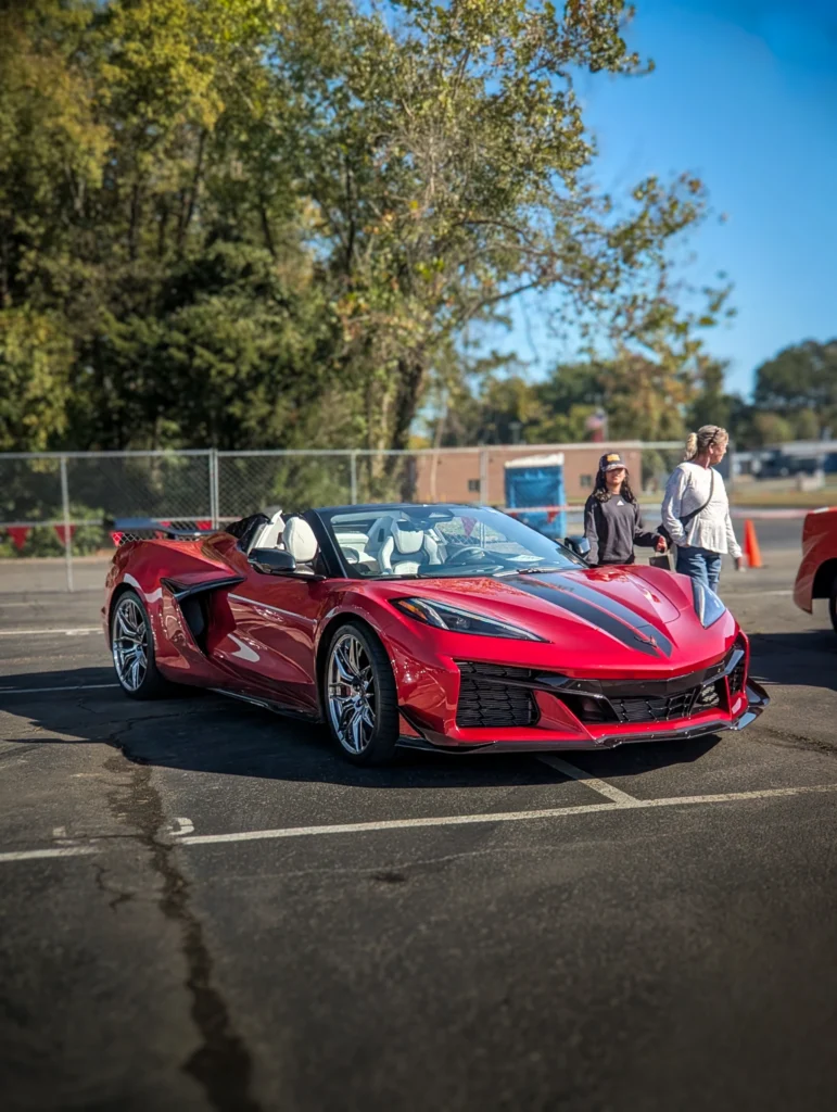 Red Chevrolet Corvette convertible parked outdoors in a lot with two people walking nearby on a sunny day