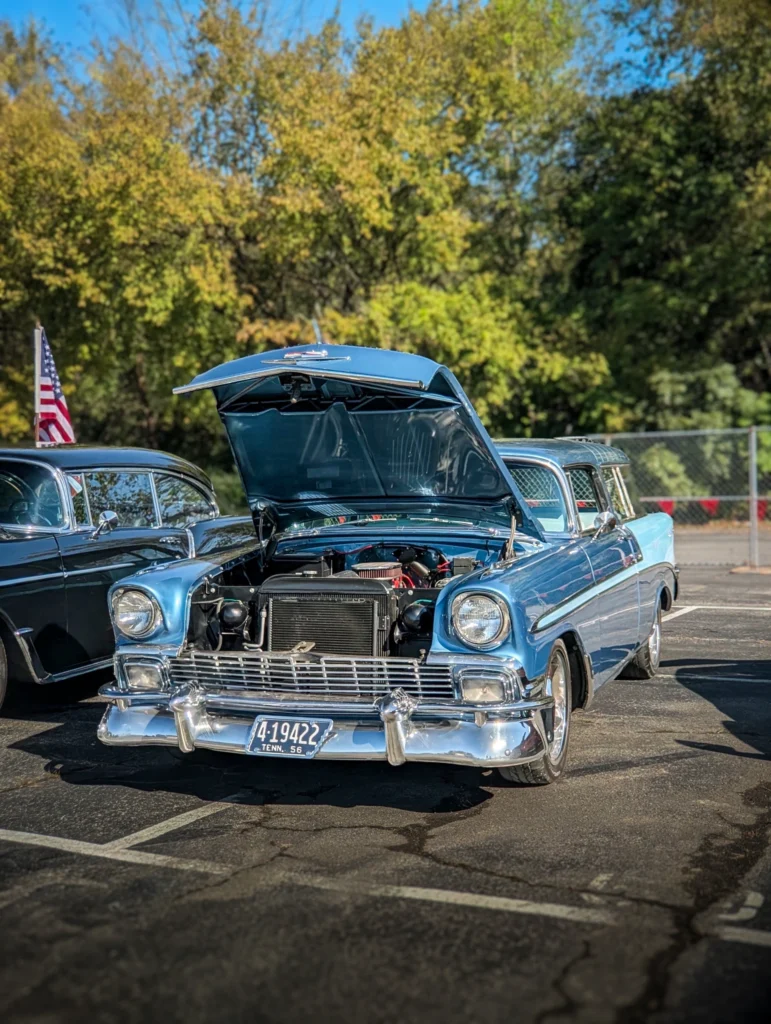 A blue Chevrolet Bel Air sits with its hood raised in a paved car show lot beside another car.