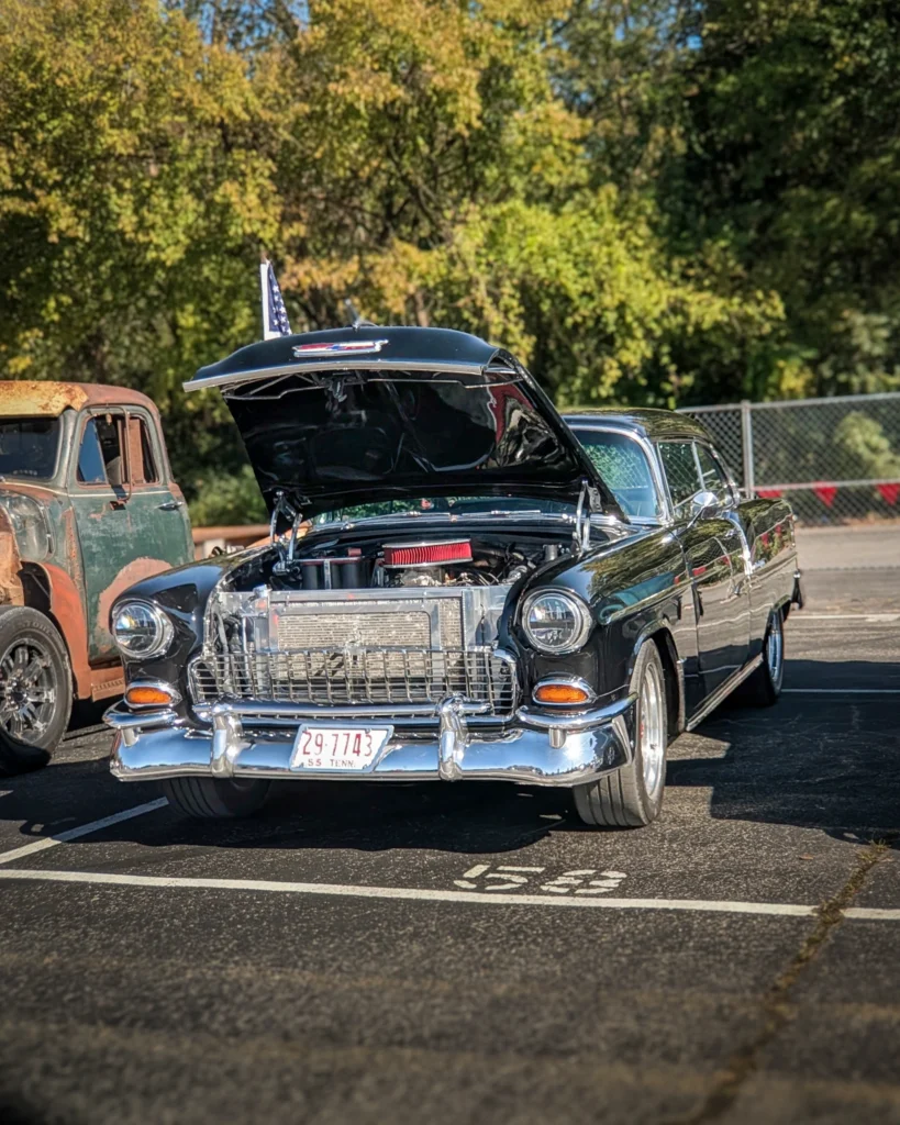 A black Chevrolet Bel Air sits with its hood raised in an outdoor parking lot beside a weathered pickup.