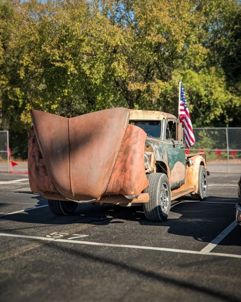 Rusty Chevrolet truck with open hood and American flag in parking lot with trees behind