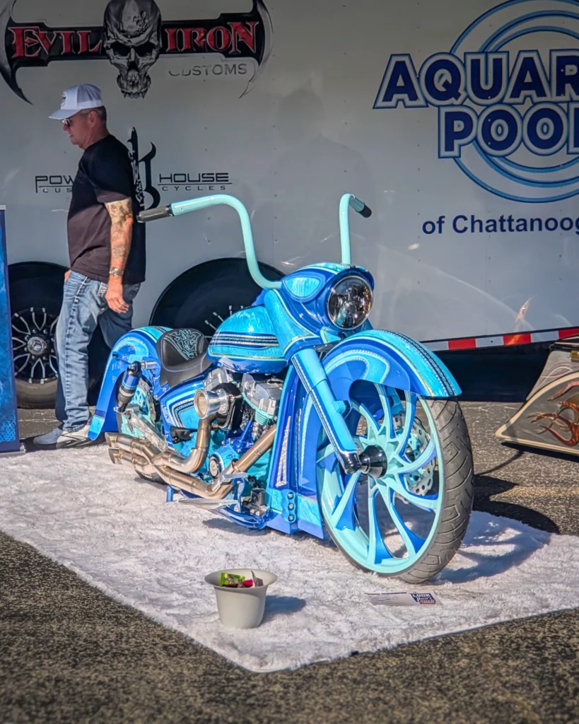 Custom blue motorcycle with extended handlebars parked on a white mat beside a man in sunglasses and cap near a trailer
