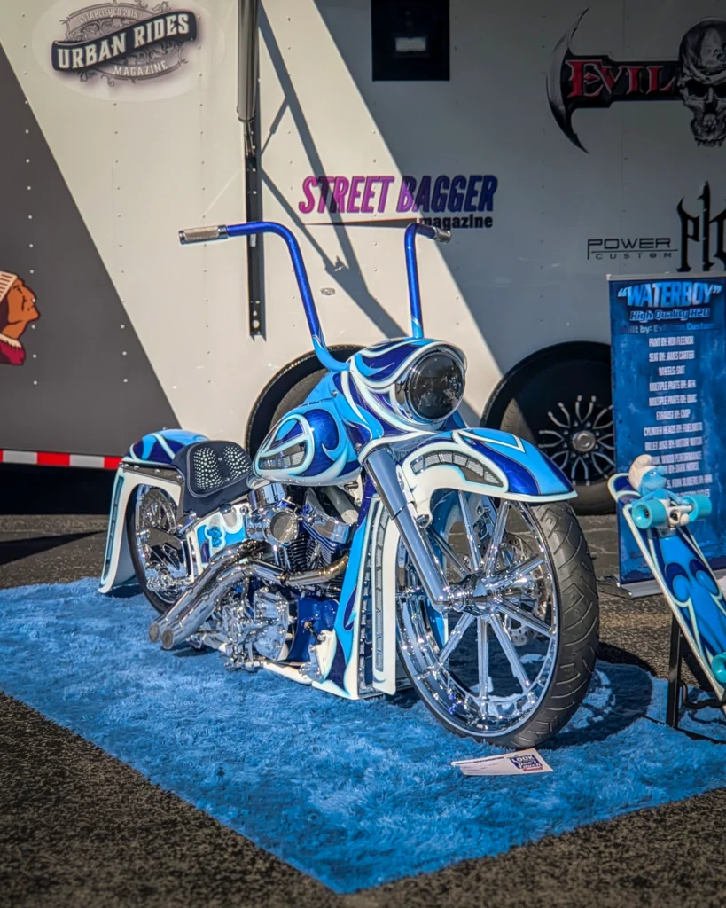 Blue and white custom motorcycle with high handlebars on a blue carpet in front of a trailer with logos and signage.