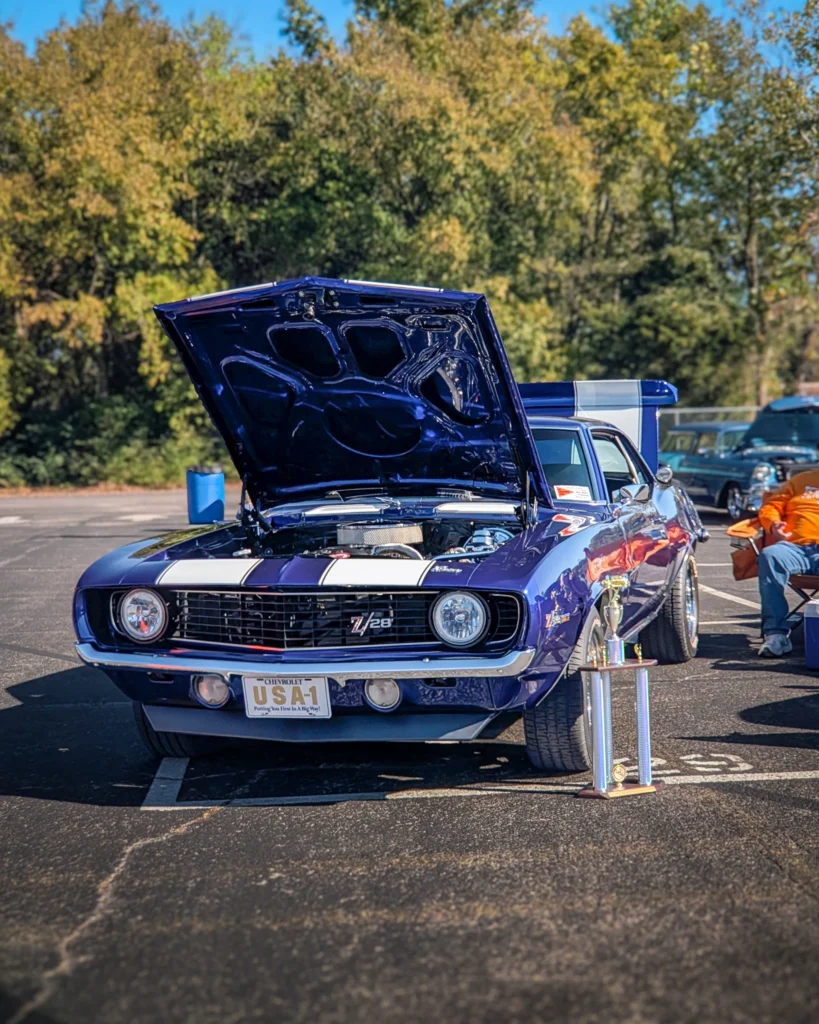 Blue Chevrolet Camaro Z28 with white racing stripes and hood open at an outdoor car show with trophies in front