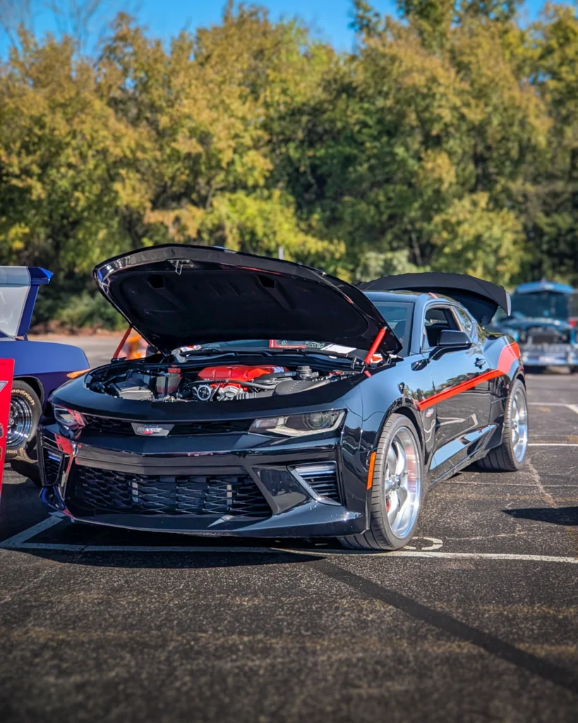 Black Chevrolet Camaro with hood open showing engine at an outdoor car event with trees in background