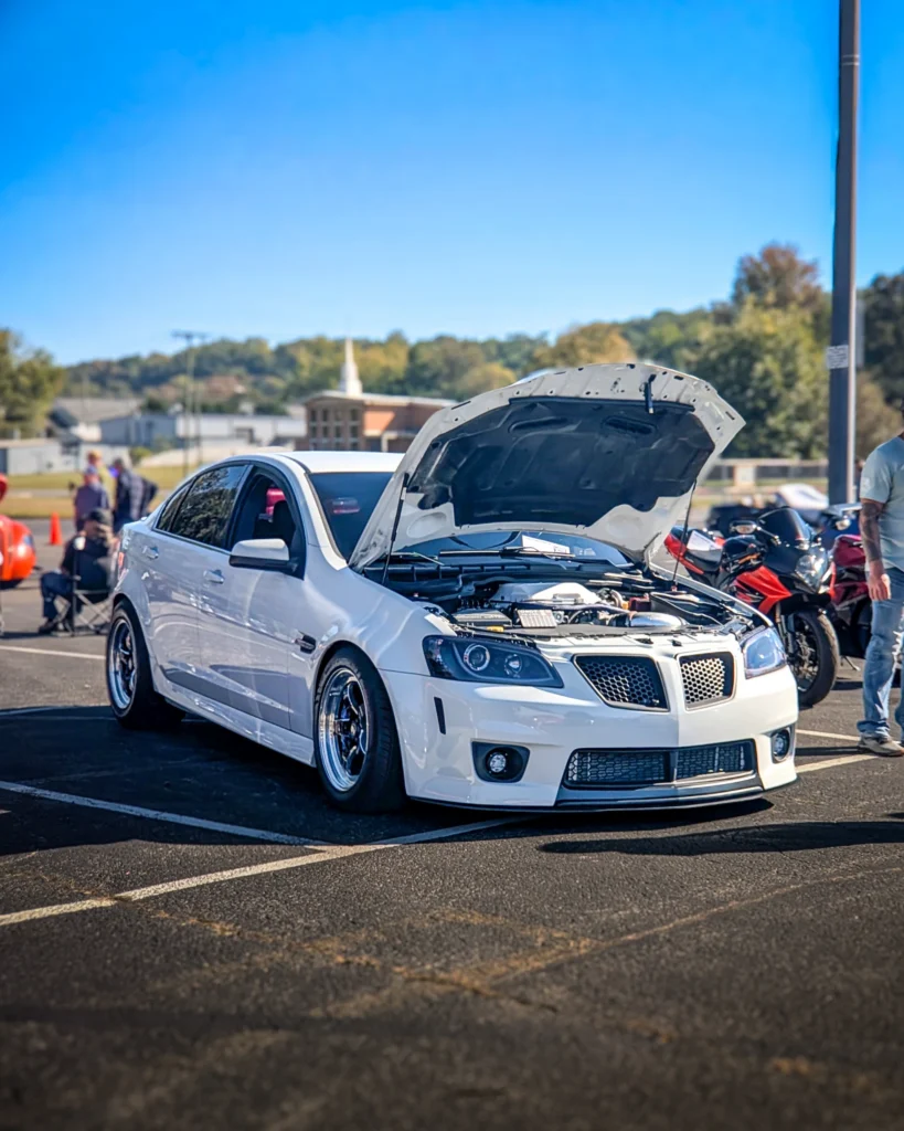 White Pontiac G8 sedan with open hood at a car event on a sunny day in a parking lot
