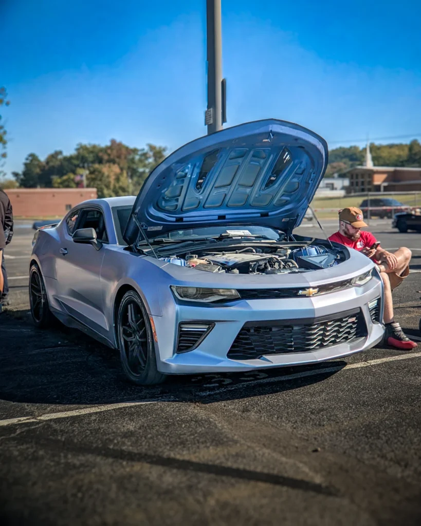 Silver Chevrolet Camaro with hood open parked outside on a sunny day near people sitting and trees in background