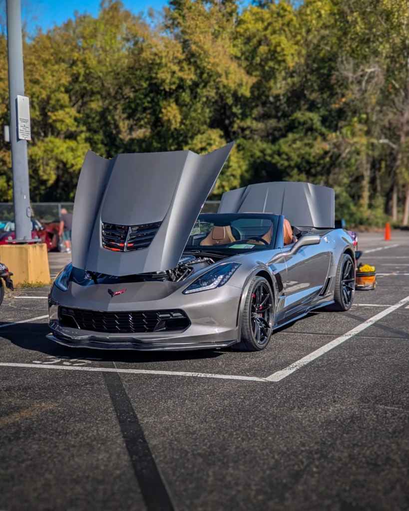 Silver Chevrolet Corvette convertible with raised hood and trunk in a parking lot at a car event