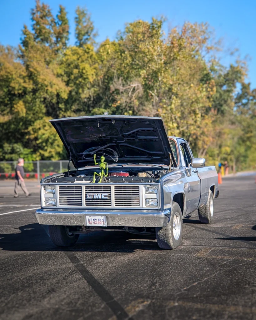 Silver GMC C/K pickup truck with open hood showing engine bay and a green skeleton figure on the grille in a parking lot