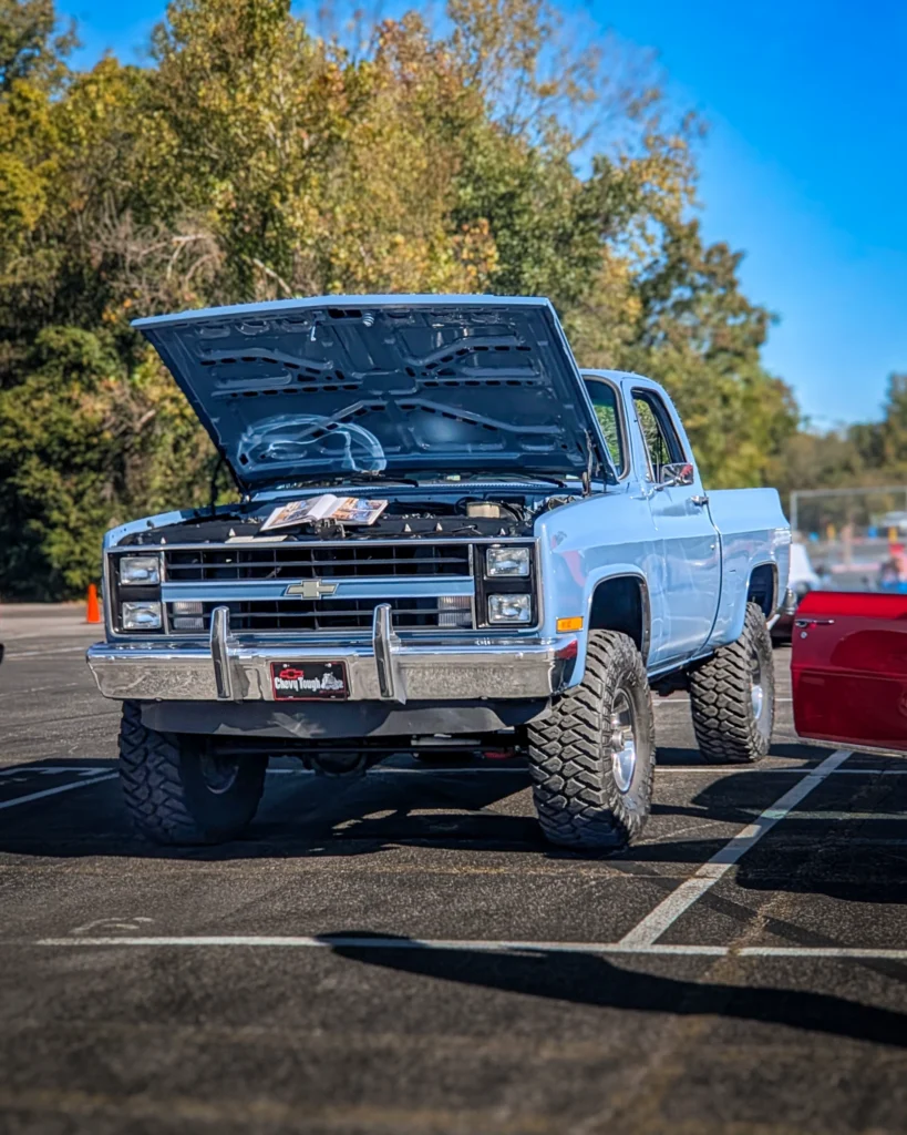 Light blue Chevrolet C/K pickup truck with raised hood in a parking lot at a car event, trees in background.