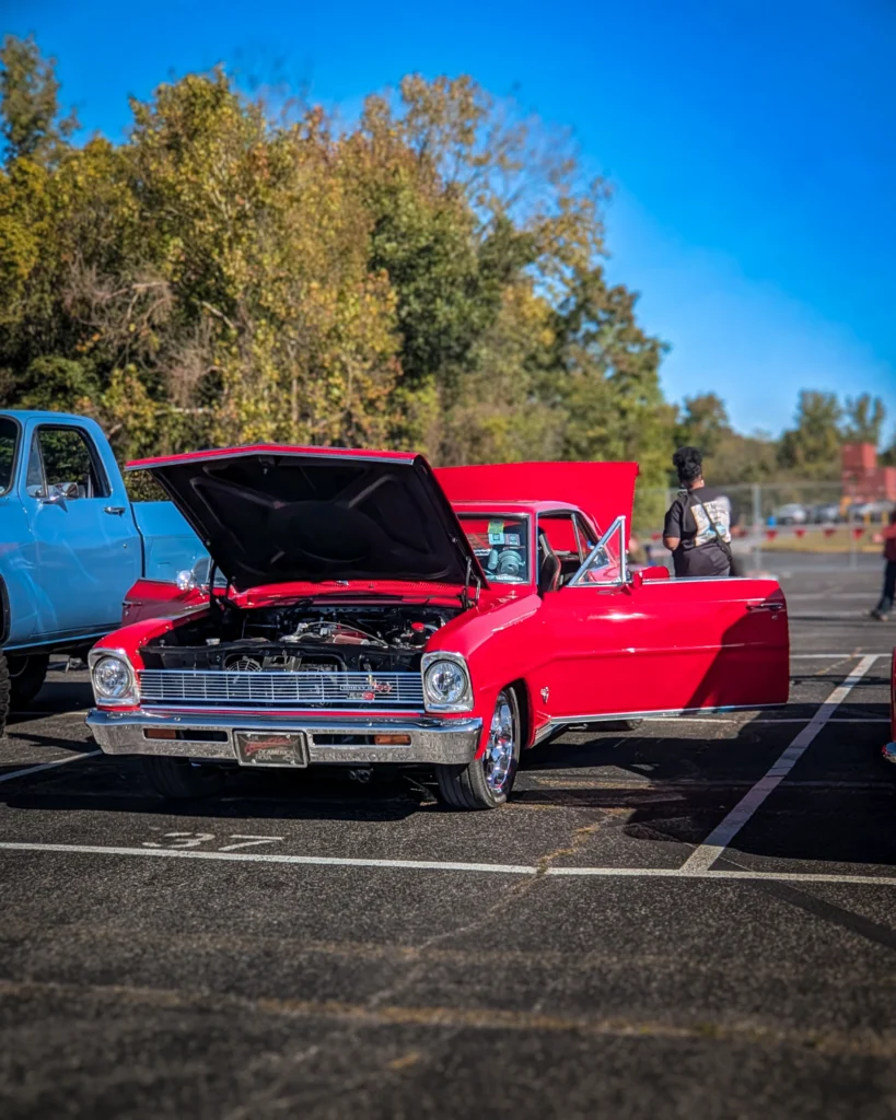 Red Chevrolet Nova SS with hood open and driver-side door open in a parking lot at a car event during daytime