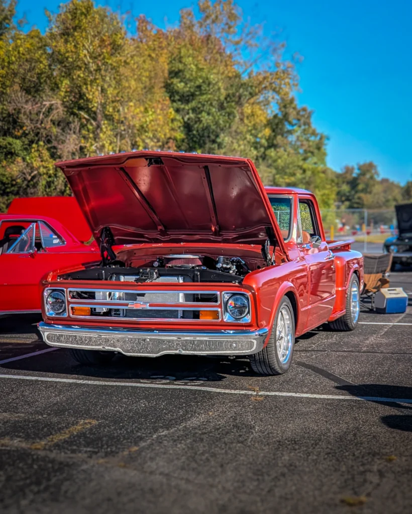 A red Chevrolet C10 pickup sits with its hood open in a parking lot at an outdoor car show.