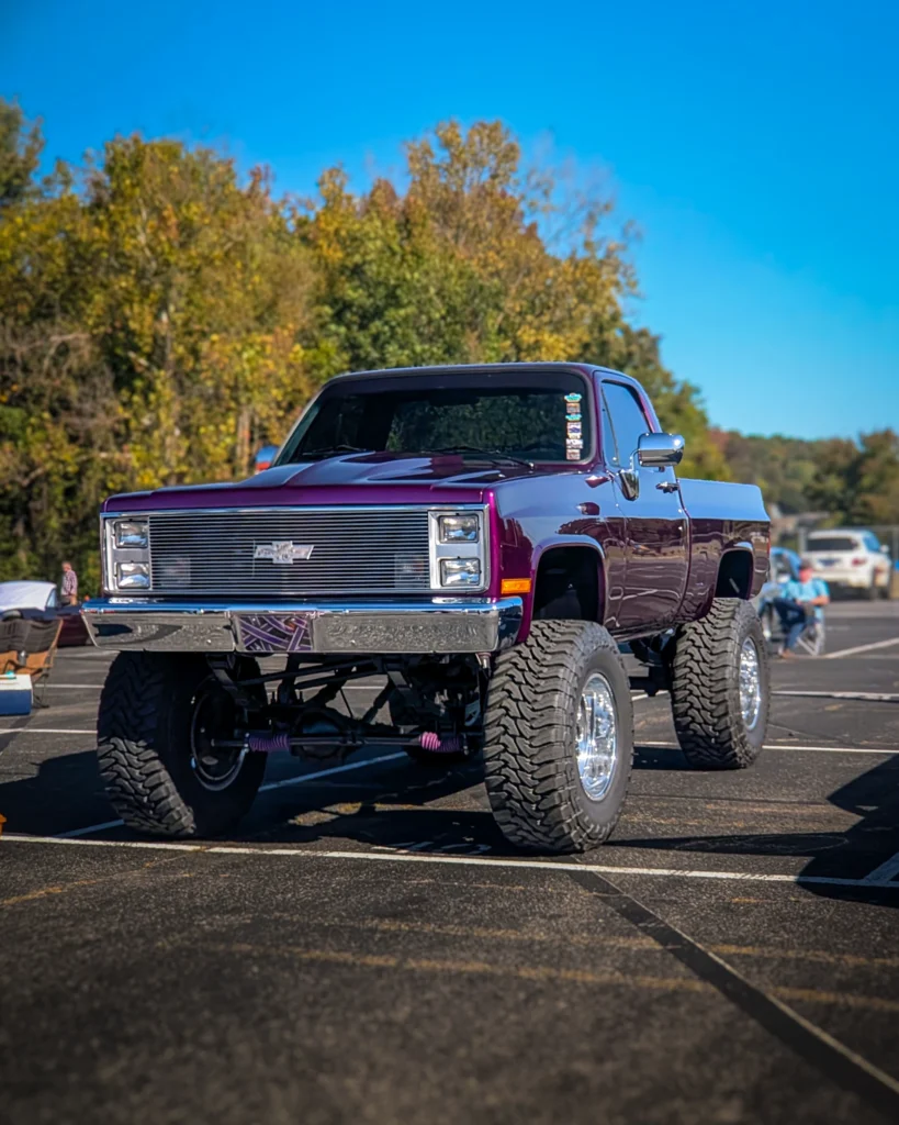 Purple lifted Chevrolet C/K pickup truck parked in an outdoor lot with trees and people in the background
