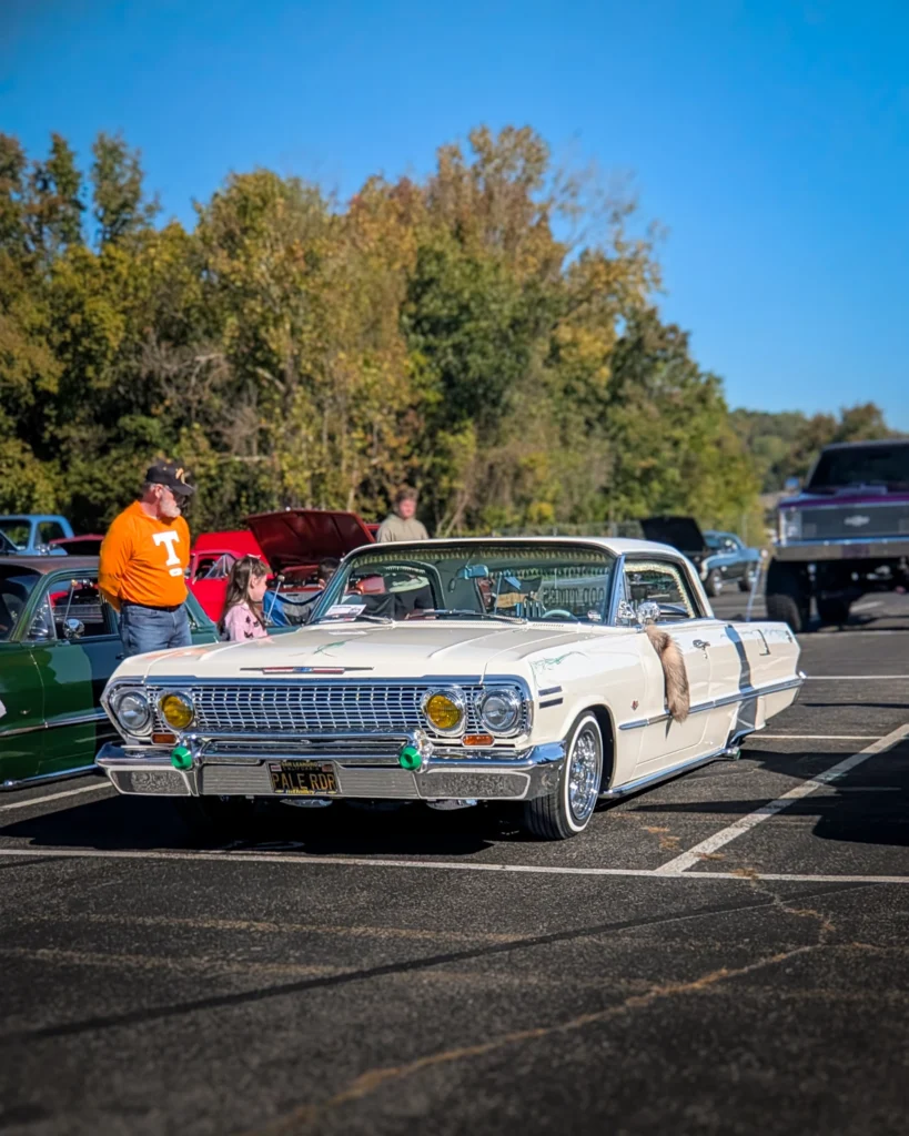 White Chevrolet Impala convertible with pinstriping parked outdoors at a car event with people nearby and trees in the
